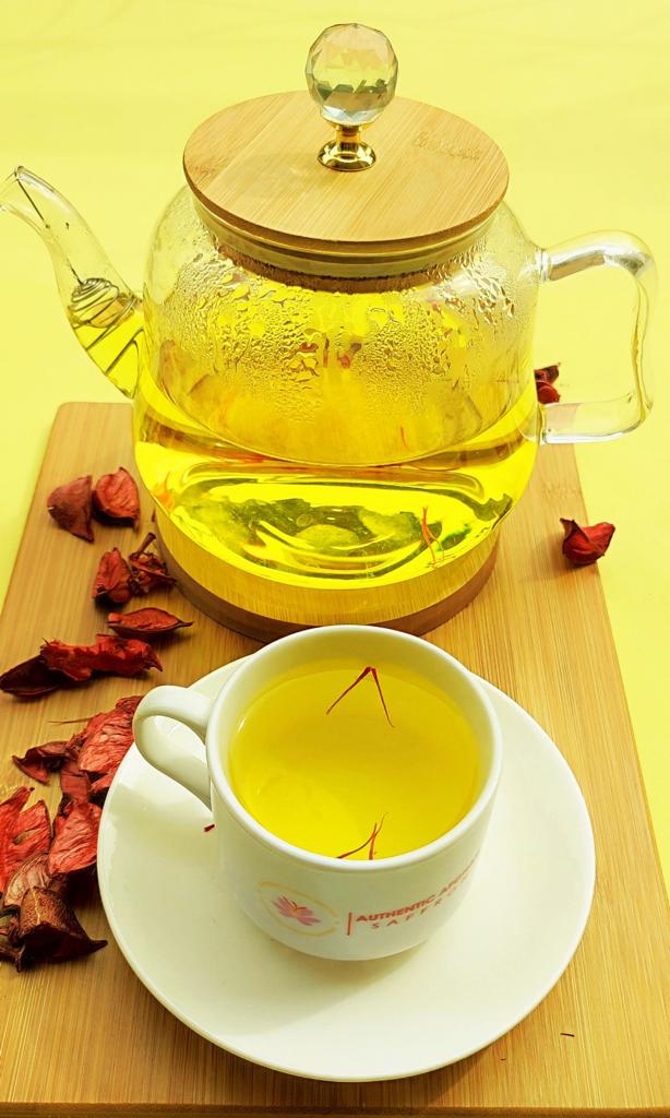 A glass teapot of golden Afghan saffron tea with a branded teacup on a bamboo board