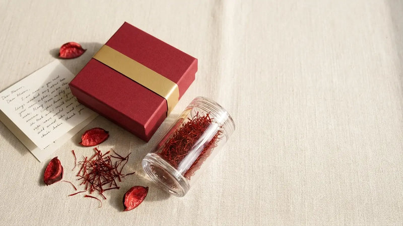 A deep red saffron gift box with a sealed crystal jar of threads and a handwritten card on linen