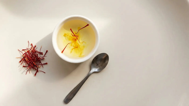 A measured pinch of saffron threads beside a small bowl of warm water where threads are blooming gold