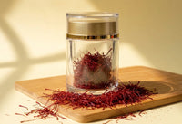 One gram of Authentic Afghan Super Negin saffron in its crystal jar on a bamboo board, with loose all-red Herati threads scattered around the base