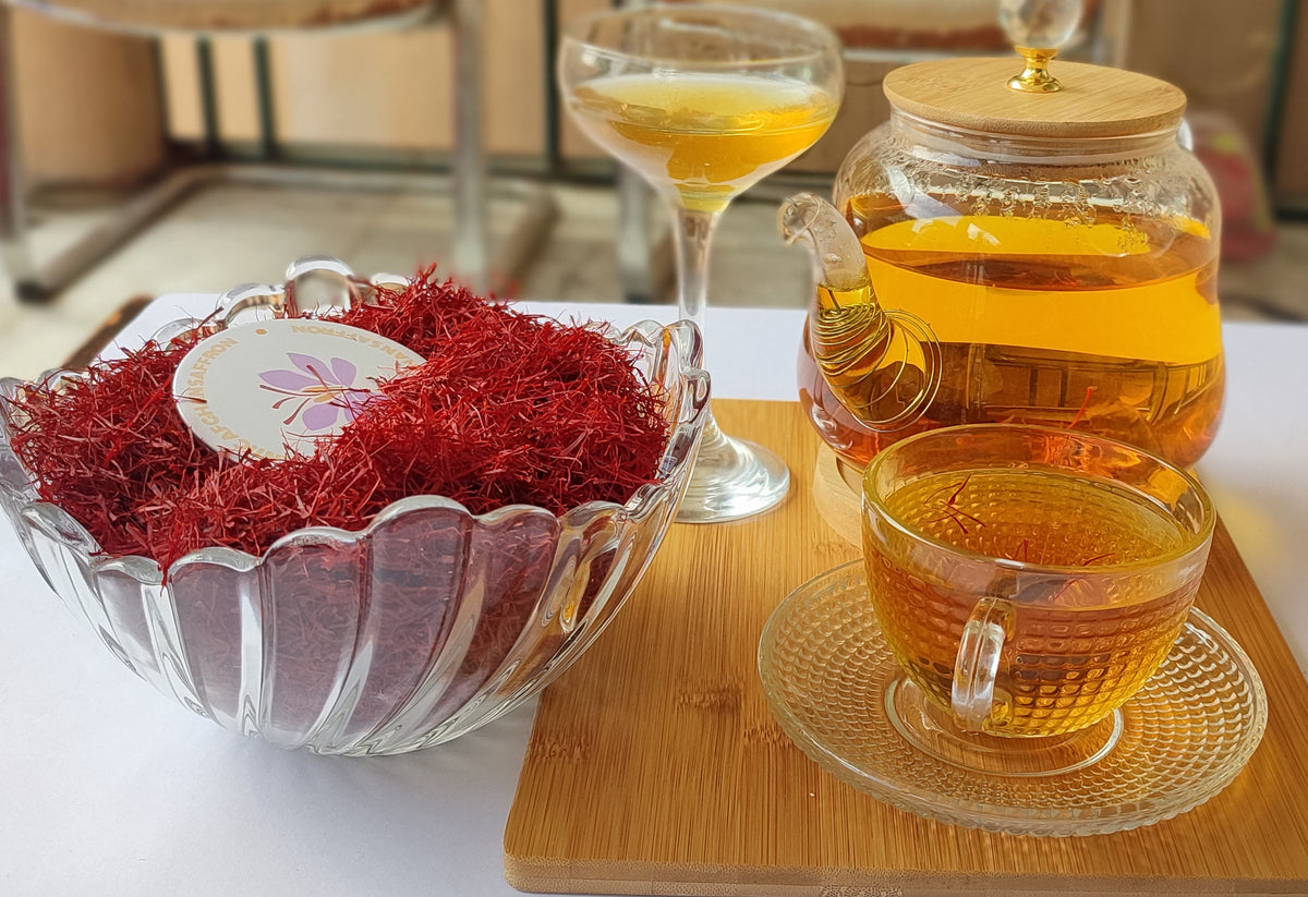 A crystal bowl heaped with all-red Afghan Super Negin saffron threads beside a glass teapot and cup of freshly brewed golden saffron tea on a bamboo board
